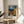 May include: Framed Van Gogh-style poster displaying a Mediterranean village scene with swirling blue night sky, golden moon, and warm yellow buildings with terracotta roofs. The print sits on a light wood round side table next to a beige upholstered chair, with natural sunlight casting shadows across white walls and a plant visible in the corner.