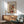 May include: Abstract watercolor artwork by Kandinsky displayed in thin wood frame on white wall above wooden sideboard. Painting features flowing organic shapes in orange, green, blue and black with curved lines and circular forms. Mid-century modern living room setting with sage green upholstered chair, white ceramic table lamp, and natural sunlight casting shadows across the scene.