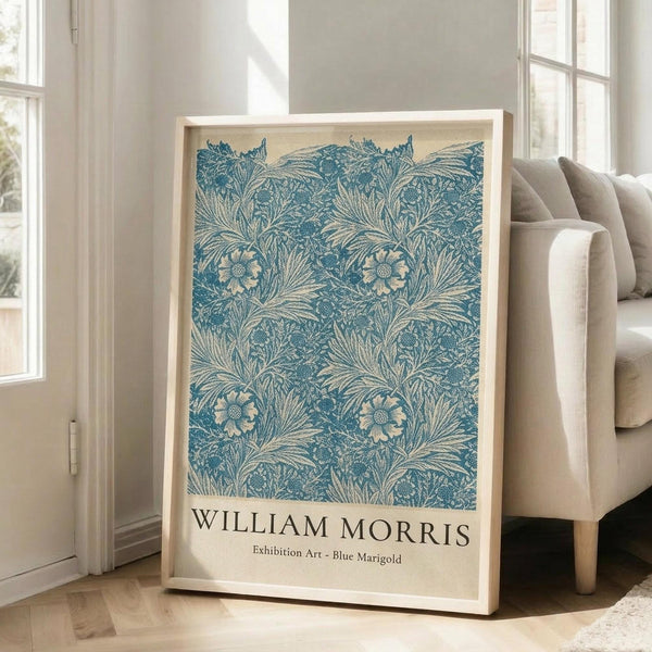 May include: Large framed poster with blue floral pattern featuring intricate botanical design of marigolds and leaves on cream background, displayed in bright white room setting next to beige upholstered chair and tall windows with natural lighting casting shadows on hardwood floors.