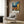 May include: Vincent van Gogh inspired poster print hanging on wood-paneled wall above beige upholstered armchair with geometric pillow. Artwork shows swirling night sky with bright moon over Mediterranean village with orange rooftops, church spire, and yellow buildings. Modern living room setting with brass floor lamp, black side table with open book, and light wood flooring.