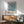 May include: Blue and cream botanical pattern poster with "WILLIAM MORRIS" text displayed in wooden frame on white wall above natural wood sideboard. Mid-century modern sage green upholstered chair with wooden arms sits beside small round table. White ceramic table lamp and bright natural lighting complete the minimalist room scene.