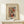 May include: Framed abstract watercolor art print in light wood frame, displayed on pale wooden console table against cream wall. Artwork features flowing organic shapes in orange, green, blue and black with curved lines. Small white wrapped gift with evergreen sprig sits beside frame. Soft natural lighting creates gentle shadows.