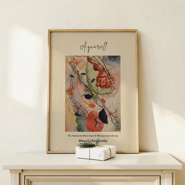 May include: Framed abstract watercolor art print in light wood frame, displayed on pale wooden console table against cream wall. Artwork features flowing organic shapes in orange, green, blue and black with curved lines. Small white wrapped gift with evergreen sprig sits beside frame. Soft natural lighting creates gentle shadows.