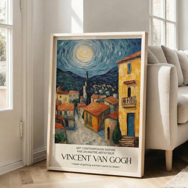 May include: Framed art print displayed against beige sofa in bright living room with white walls and large windows. Poster features Van Gogh-inspired painting of Mediterranean village with orange terracotta roofs under swirling starry night sky. Yellow and blue buildings line cobblestone streets beneath luminous moon. French text and artist name printed below artwork in white border. Natural wood frame, approximately 2-3 feet tall.