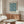 May include: Framed poster with blue floral marigold pattern hung above wooden sideboard in minimalist living room. White curved chair and glass-top coffee table visible. Poster features intricate blue botanical design on cream background in thin wooden frame. Modern Scandinavian-style interior with neutral walls and natural wood furniture.