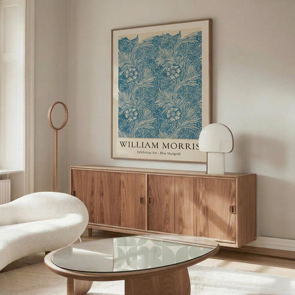 May include: Framed poster with blue floral marigold pattern hung above wooden sideboard in minimalist living room. White curved chair and glass-top coffee table visible. Poster features intricate blue botanical design on cream background in thin wooden frame. Modern Scandinavian-style interior with neutral walls and natural wood furniture.