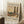 May include: Framed art print featuring a colorful landscape painting with two tall poplar trees, rural buildings, and winding path in earthy tones of green, orange, and brown. The poster displays exhibition text from The Art Institute of Chicago for Wassily Kandinsky. Frame leans against white paneled wall on textured cream rug, with wooden side table holding white coffee cup and saucer, and beige throw blanket visible in corner.