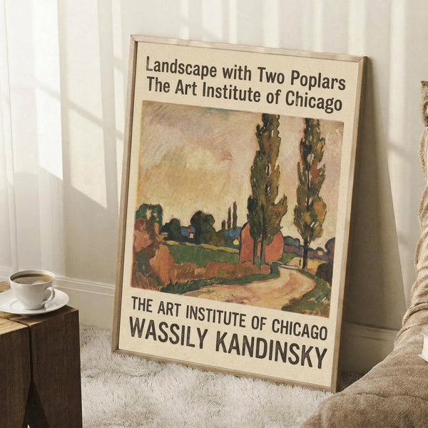 May include: Framed art print featuring a colorful landscape painting with two tall poplar trees, rural buildings, and winding path in earthy tones of green, orange, and brown. The poster displays exhibition text from The Art Institute of Chicago for Wassily Kandinsky. Frame leans against white paneled wall on textured cream rug, with wooden side table holding white coffee cup and saucer, and beige throw blanket visible in corner.