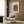 May include: Beige framed poster with abstract geometric shapes in purple and cream tones hanging on wood-paneled wall above light gray upholstered armchair with geometric patterned pillow. Modern brass floor lamp and dark round side table with open book nearby. Warm neutral interior with natural lighting from window.