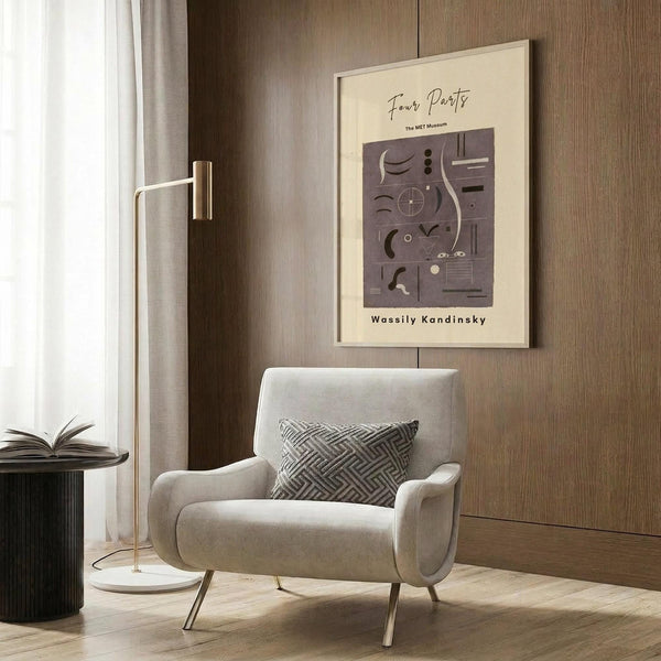 May include: Beige framed poster with abstract geometric shapes in purple and cream tones hanging on wood-paneled wall above light gray upholstered armchair with geometric patterned pillow. Modern brass floor lamp and dark round side table with open book nearby. Warm neutral interior with natural lighting from window.
