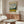 May include: Van Gogh museum poster featuring Wheat Field with Cypresses painting displayed above wooden credenza in modern living room. Poster shows swirling blue sky, golden wheat field, and dark cypress tree. White sculptural chair and glass coffee table visible. Warm wood tones throughout minimalist space with cream walls.