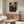 May include: Framed poster featuring a blue bird perched among ornate floral patterns with red roses and cream flowers on dark teal background, displayed above mid-century walnut credenza in minimalist living room with white upholstered chair, glass oval coffee table, and modern floor lamp