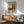 May include: Framed Kandinsky landscape poster with two tall poplar trees, colorful fields, and yellow sky hanging above wood credenza with white ceramic lamp. Mid-century modern green upholstered chair with wooden arms sits nearby on hardwood floor. Bright natural light creates geometric shadows on white wall in minimalist living room setting.