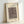 May include: Framed abstract art print leaning against white wall corner. Light wood frame contains cream poster with gray abstract geometric composition featuring curved lines, circles, rectangles and various shapes in muted tones. Artist name printed below artwork in simple black text.
