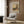 May include: Framed vintage art exhibition poster featuring Wassily Kandinsky's colorful landscape painting with two tall poplar trees, winding path, and buildings. Displayed on wood-paneled wall above beige upholstered armchair with geometric patterned pillow, brass floor lamp, and small round side table with open book in modern living room setting.