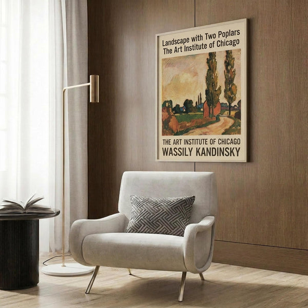 May include: Framed vintage art exhibition poster featuring Wassily Kandinsky's colorful landscape painting with two tall poplar trees, winding path, and buildings. Displayed on wood-paneled wall above beige upholstered armchair with geometric patterned pillow, brass floor lamp, and small round side table with open book in modern living room setting.