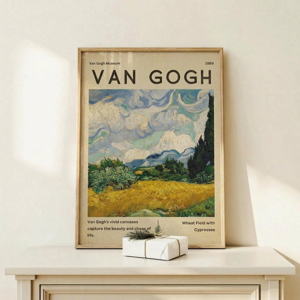 May include: Framed Van Gogh museum poster featuring Wheat Field with Cypresses painting, showing swirling blue sky over golden wheat field with green cypress trees and mountains. Poster has cream background with black text and 1889 date. Natural wood frame sits on light wooden surface against white wall, with small white wrapped gift box with twine and evergreen sprig beside it.