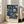 May include: Framed poster with intricate blue floral pattern on dark background featuring ornate blooms and swirling foliage in William Morris decorative style, displayed in light wood frame leaning against white wall next to beige upholstered sofa in bright, airy living room with large windows