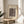 May include: Abstract art print in wooden frame featuring geometric shapes and lines in brown tones on cream background, displayed on round wooden side table next to beige upholstered chair with decorative pillow, green plant leaves visible in corner, natural sunlight casting shadows across neutral-toned room setting.