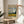 May include: Van Gogh museum poster featuring "Wheat Field with Cypresses" painting displayed in thin wooden frame on round wooden stool. Person in beige sweater holds poster showing swirling blue sky, golden wheat field, dark cypress trees, and green landscape. Sunlit room with white walls, plant leaves, and neutral furniture visible.