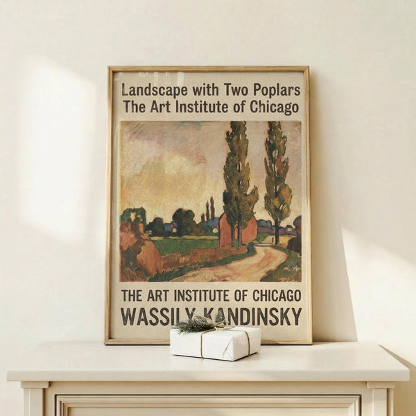 May include: Framed poster showing Kandinsky's landscape painting with two tall poplar trees, winding dirt path, and rural buildings in earthy tones of green, brown, and orange. Exhibition poster format with "The Art Institute of Chicago" text. Light wood frame leaning against white wall on pale wooden surface with small wrapped gift box nearby.