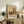 May include: Framed art print featuring a colorful landscape painting with two tall poplar trees, winding path, and buildings in warm oranges and greens, displayed on a rustic wooden sideboard alongside potted plants, books, and ceramic vases in a cozy living room setting with neutral tones and natural lighting.