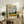 May include: Framed Van Gogh museum poster featuring swirling blue and white clouds above a golden wheat field with dark cypress trees. The vintage-style print sits on a rustic wooden sideboard alongside ferns, books, and ceramic pottery in a neutral-toned living room setting with a cream upholstered chair.