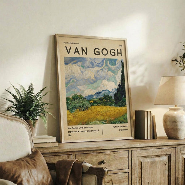May include: Framed Van Gogh museum poster featuring swirling blue and white clouds above a golden wheat field with dark cypress trees. The vintage-style print sits on a rustic wooden sideboard alongside ferns, books, and ceramic pottery in a neutral-toned living room setting with a cream upholstered chair.