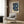 May include: Framed William Morris floral art print with blue flowers and cream botanical motifs hanging on wood paneled wall above beige upholstered armchair with geometric patterned pillow, gold floor lamp, and dark round side table with open book in modern living room setting