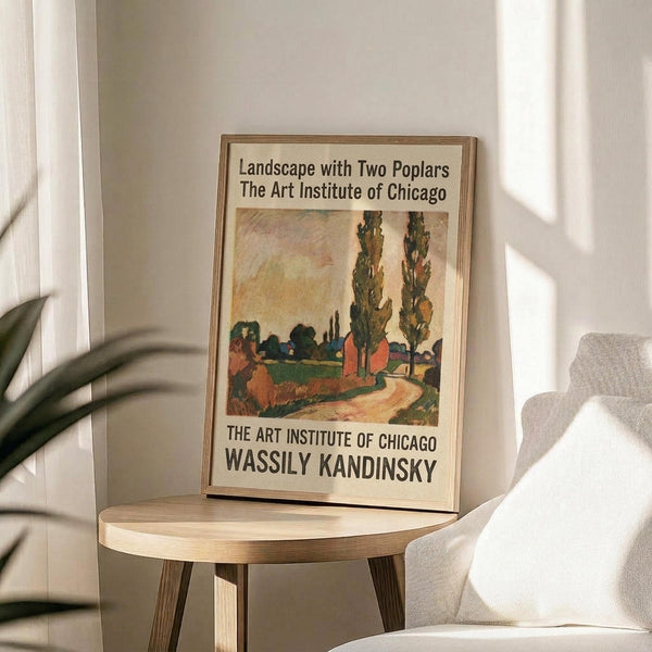 May include: Framed poster print featuring a colorful landscape painting with two tall poplar trees, winding path, and vibrant autumn colors in oranges, greens, and yellows. The print includes museum text identifying it as a Wassily Kandinsky work from The Art Institute of Chicago. Displayed on a light wood round side table next to a beige upholstered chair with plant leaves visible in soft natural lighting.