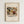 May include: Framed art print mounted on white textured wall showing Kandinsky's abstract expressionist painting with flowing organic forms in earth tones of brown, green, yellow and red, plus black linear elements. Natural wood frame surrounds cream-colored mat with artwork title and date below the image.