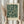 May include: Hands holding a framed botanical art print featuring an intricate dark green and cream floral pattern with flowing leaves and small blossoms in William Morris style. The print is displayed in a light wood frame against soft curtains with dried pampas grass visible to the side, creating a cozy neutral-toned interior setting.