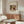 May include: Abstract expressionist artwork poster in wooden frame hanging above walnut credenza in modern living room. Colorful abstract painting features flowing organic shapes in earth tones - browns, oranges, greens, and blacks with dynamic brushstrokes. Room styled with cream upholstered chair, glass oval coffee table, and white sculptural table lamp on sideboard.