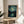 May include: Framed poster print of a swirling night sky with bright stars and crescent moon over a village reflected in water below, displayed on a round wooden side table next to a beige upholstered chair. Sunlight streams across white walls creating geometric shadows, with a green plant visible in the corner. The artwork features deep blues and yellows in an impressionist style with visible brushstroke textures.
