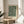 May include: Framed botanical art print with dark green background and cream-colored flowing leaves and small flowers in William Morris style, displayed on light wood round side table next to beige upholstered chair, with plant leaves visible in corner and natural sunlight streaming across white walls.