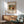 May include: Abstract expressionist artwork poster in wooden frame hanging above mid-century modern wooden credenza with white table lamp. Green upholstered armchair with wooden arms sits nearby. The colorful abstract painting features swirling forms in yellow, orange, red, green and black on cream background. Natural sunlight streams across white wall creating geometric shadows in minimalist living room setting.