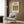 May include: Cream-colored framed poster featuring vintage angel illustration hanging on wood-paneled wall above beige upholstered armchair with geometric patterned pillow. Modern floor lamp with brass finish and round side table with open book complete the neutral-toned reading nook scene.