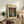 May include: Framed Van Gogh poster print featuring orange poppies in white vase against blue background, displayed on rustic wood sideboard beside fern plant, books, ceramic vase and beige upholstered chair in neutral-toned living room setting