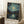 May include: Framed poster print of Van Gogh's Starry Night style artwork showing swirling night sky with glowing stars and moon over water with reflections and small boat. Blue and yellow dominant colors with thick brushstroke texture. Natural wood frame leaning against white wall corner next to wooden side table with white coffee cup and saucer, cream textured rug below.