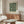 May include: Dark green botanical poster with cream-colored leaves and flowers in Arts and Crafts style, displayed in thin wooden frame above mid-century modern walnut credenza. Room features white upholstered chair, glass-top coffee table, and minimalist decor with natural lighting.