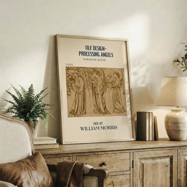May include: Framed vintage William Morris angel artwork print displayed on rustic wooden sideboard in neutral-toned living room. Brown sepia-toned illustration shows three robed angels in medieval style. Cream matting in light wood frame, surrounded by fern plant, ceramic vase, books, and beige upholstered chair with leather throw pillow.