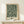 May include: Framed botanical poster with dark green background and cream-colored flowing floral pattern, displayed on light wooden console table against white wall. Small wrapped gift box with evergreen sprig sits beside the natural wood frame. Soft natural lighting creates gentle shadows on the minimalist scene.
