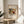 May include: Framed art print of abstract expressionist painting with colorful organic shapes in blues, greens, yellows, and reds against cream background. Print displays on round wooden side table next to beige upholstered chair. Natural sunlight creates soft shadows across white wall. Green plant leaves visible in corner. Warm, minimalist interior styling.