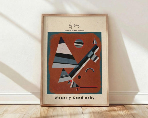 May include: Framed abstract art print in light wood frame leaning against white wall on wooden floor. Artwork features geometric shapes in rust red, teal blue, black and white - triangular forms, circles, and angular lines in modernist style. Cream matting surrounds the colorful composition. Natural lighting creates soft shadows on textured white wall.
