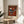 May include: Abstract modernist artwork poster in wooden frame displaying geometric shapes in rust orange, teal, black and white - triangles, circles and angular forms against burnt orange background. Museum exhibition poster with "GRIS" and "WASSILY KANDINSKY" text at bottom. Frame sits on round wooden side table next to beige upholstered chair with plant leaves visible, sunlight casting shadows on white wall.