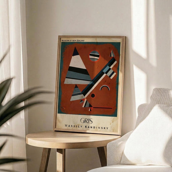 May include: Abstract modernist artwork poster in wooden frame displaying geometric shapes in rust orange, teal, black and white - triangles, circles and angular forms against burnt orange background. Museum exhibition poster with "GRIS" and "WASSILY KANDINSKY" text at bottom. Frame sits on round wooden side table next to beige upholstered chair with plant leaves visible, sunlight casting shadows on white wall.