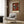 May include: Abstract art poster featuring geometric shapes in red, teal, black and white, displayed above a beige upholstered armchair with geometric pillow. Modern living room setting with brass floor lamp, dark round side table with open book, and wood panel wall. Natural lighting from window with white curtains creates warm, contemporary atmosphere.
