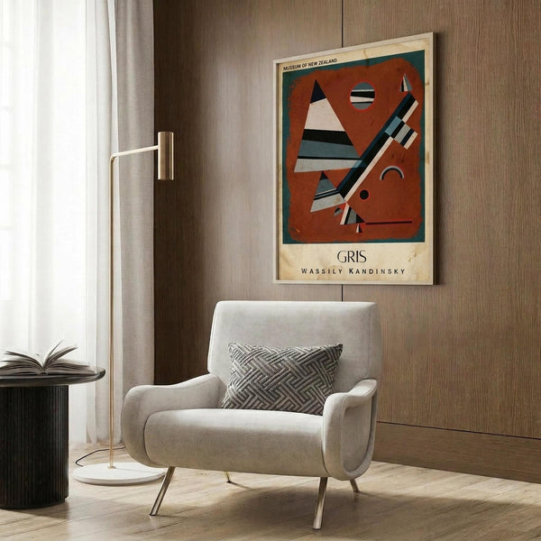 May include: Abstract art poster featuring geometric shapes in red, teal, black and white, displayed above a beige upholstered armchair with geometric pillow. Modern living room setting with brass floor lamp, dark round side table with open book, and wood panel wall. Natural lighting from window with white curtains creates warm, contemporary atmosphere.