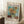 May include: Framed Van Gogh museum poster featuring three bright yellow sunflowers in a green ceramic vase against turquoise background, displayed in cozy room corner with white walls, wooden side table holding white coffee cup and saucer, cream knit throw blanket, and textured white rug