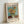 May include: Framed poster print featuring Van Gogh's sunflower painting with yellow blooms in green vase against turquoise background, displayed in light wooden frame leaning against white wall corner. Museum-style layout with "VAN GOGH" text header, artwork title "Vase with Three Sunflowers," and descriptive text about the artist's vivid canvases capturing life's beauty and chaos.